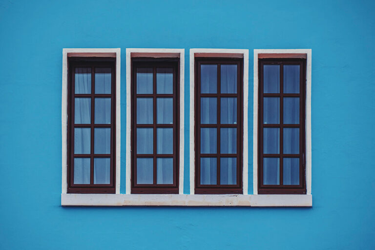 Vibrant blue stucco wall featuring four tall windows with dark wooden frames casting subtle reflections