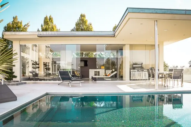 Modern white stucco home with floor-to-ceiling glass walls, reflective pool edge, loungers and minimalist patio
