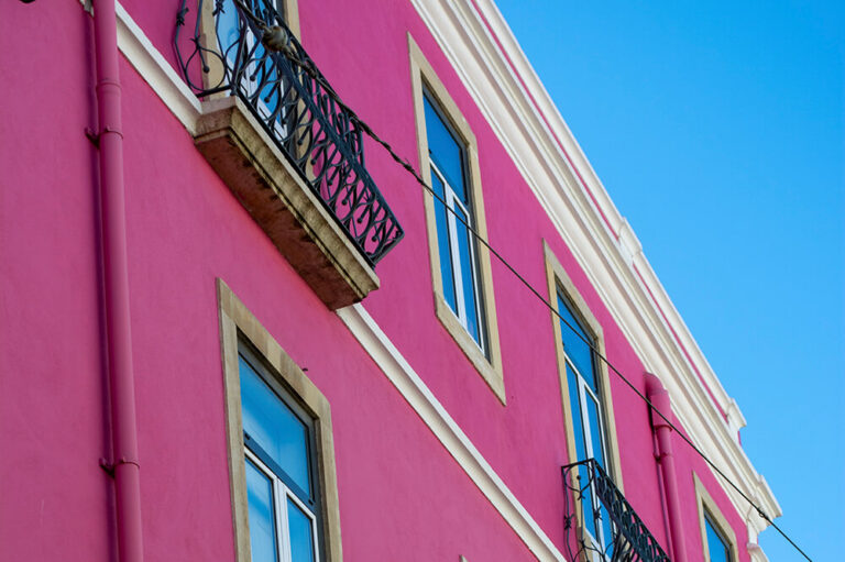Pink stucco wall with tall white-framed windows and ornate black iron balconies under clear blue sky