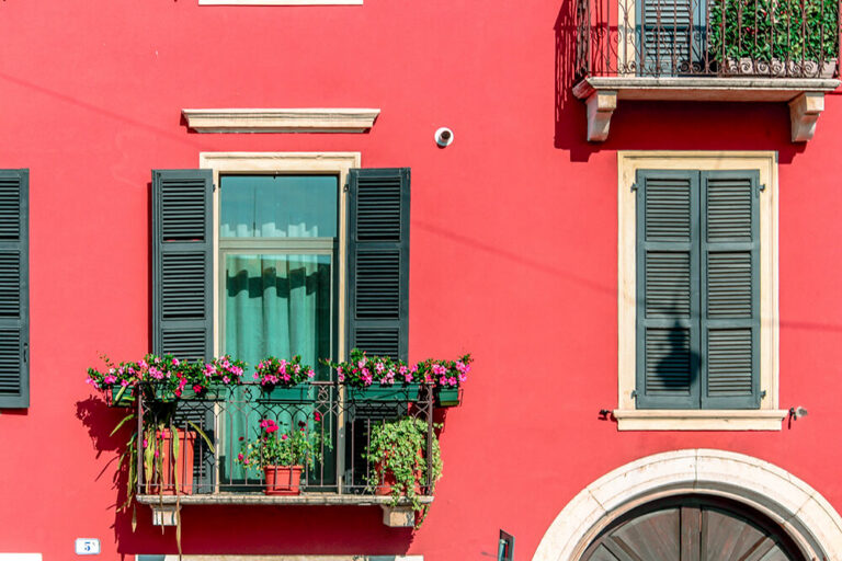 Vibrant red stucco facade with dark green shutters and a balcony adorned with pink flowering plants.