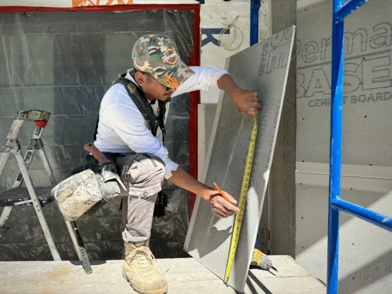 Worker measuring and marking cement board for stucco base installation on scaffold with a tape measure