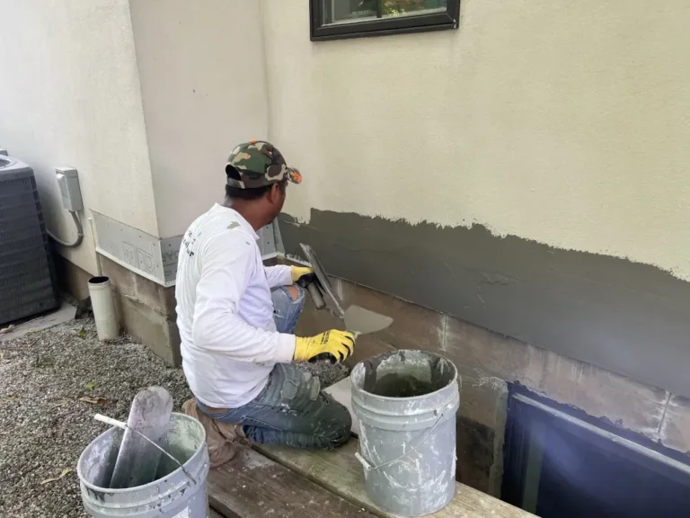Worker kneeling on bench outdoors applying gray stucco base coat to foundation wall with trowel and bucket