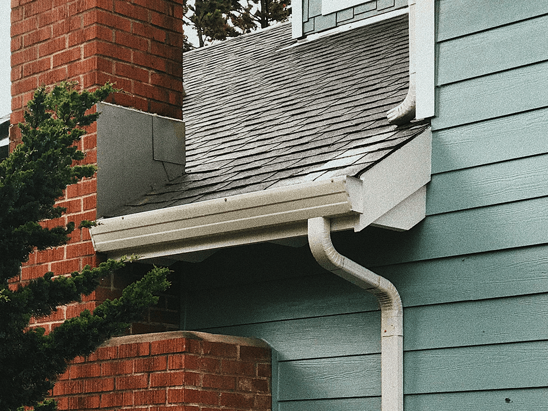 Close-up of roofline showing white gutter and downspout system attached to teal siding and red brick chimney