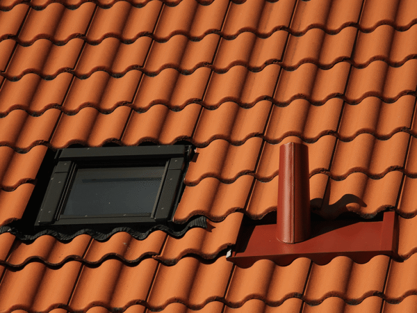 Clay Tile Roof with Skylight and Red Metal Vent Pipe Close-up of red clay tile roofing with black-framed skylight and cylindrical red vent flashing for ventilation