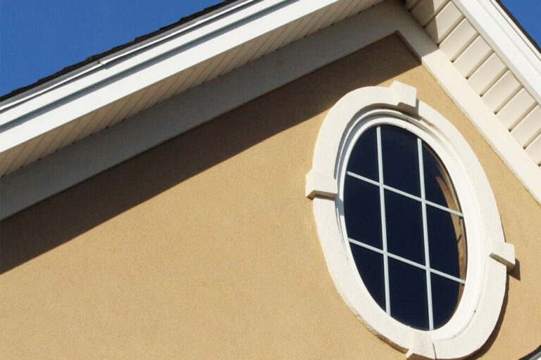 Close-up of oval window with white stucco trim on tan exterior wall under gable roof and blue sky above