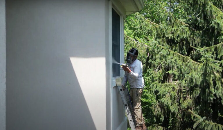 Technician on ladder sealing stucco window with spray gun, using protective gear and sealing tape for precision