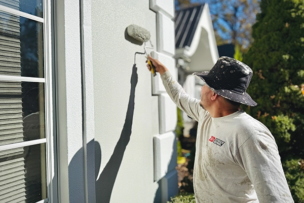 Exterior stucco painting with roller Technician applying paint to an exterior stucco wall with a roller near a window, daytime