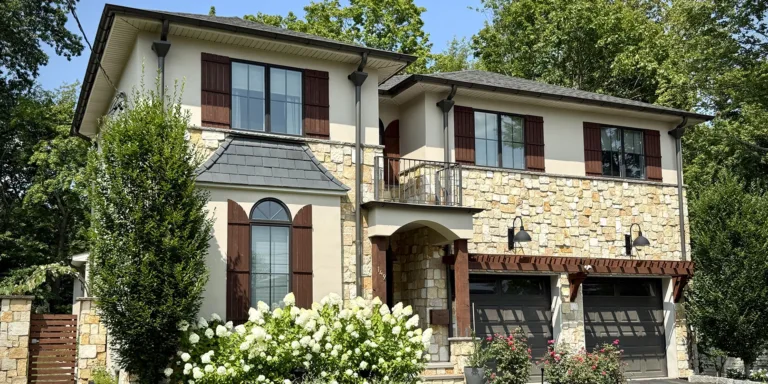 Two-story home in Paramus, NJ with light sand-finish stucco above stone veneer, arched entry, and double garage; daytime.