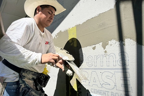 sExterior stucco installation — base coat on cement boardtucco-base-coat-on-cement-board-zd-stucco-repair Worker applying base coat and mesh on a cement board facade during stucco installation
