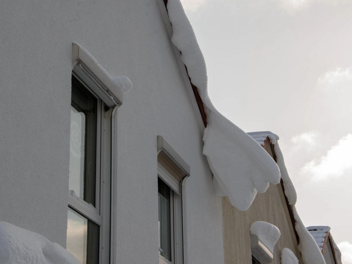 Closeup of a white stucco wall and windows with heavy roof snow hanging over the eaves in winter