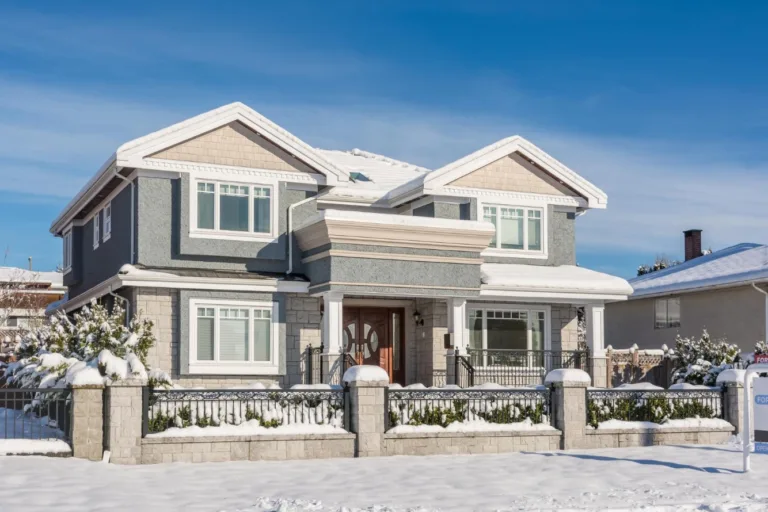 Front elevation of a gray stucco house with white trim and a snow-covered yard under a clear blue sky
