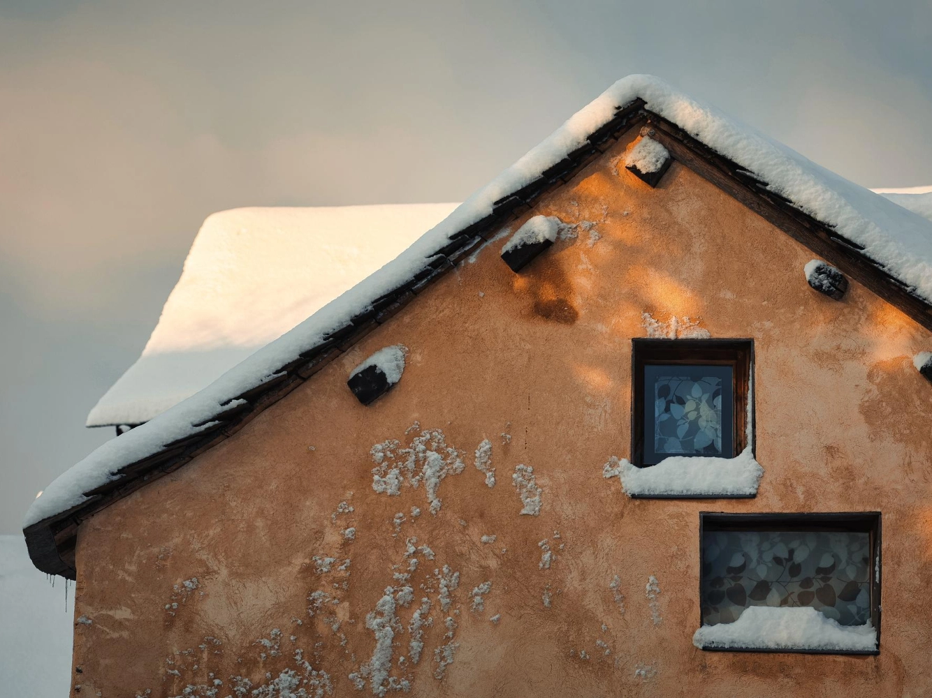 Close view of an aged stucco gable with snow on the roof and around two small windows in winter light