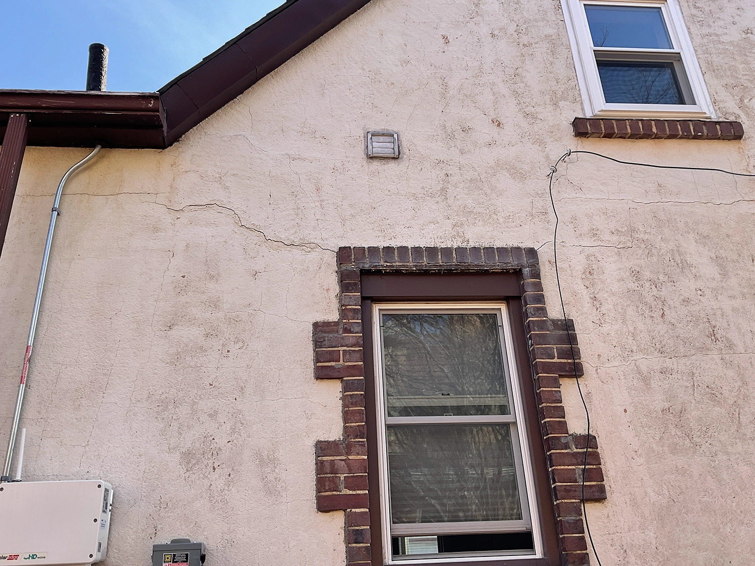 Front gable wall of an older stucco home showing long horizontal and diagonal cracks near the window and eave.