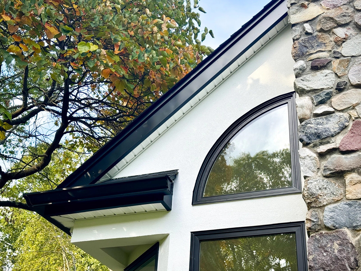 5 Common Stucco Myths Debunked – Breaking Down Popular Misconceptions 3 Close view of a white stucco gable with black trim, arched window, and adjacent natural-stone chimney under leafy tree.