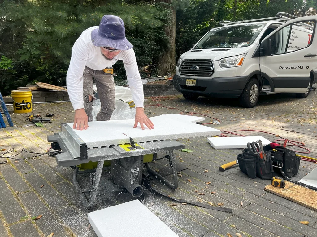 Technician cutting an EPS foam panel for EIFS stucco on a portable table saw; company van and tools visible in a driveway.