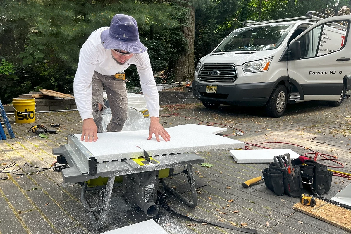 Technician cutting an EPS foam panel for EIFS stucco on a portable table saw; company van and tools visible in a driveway.