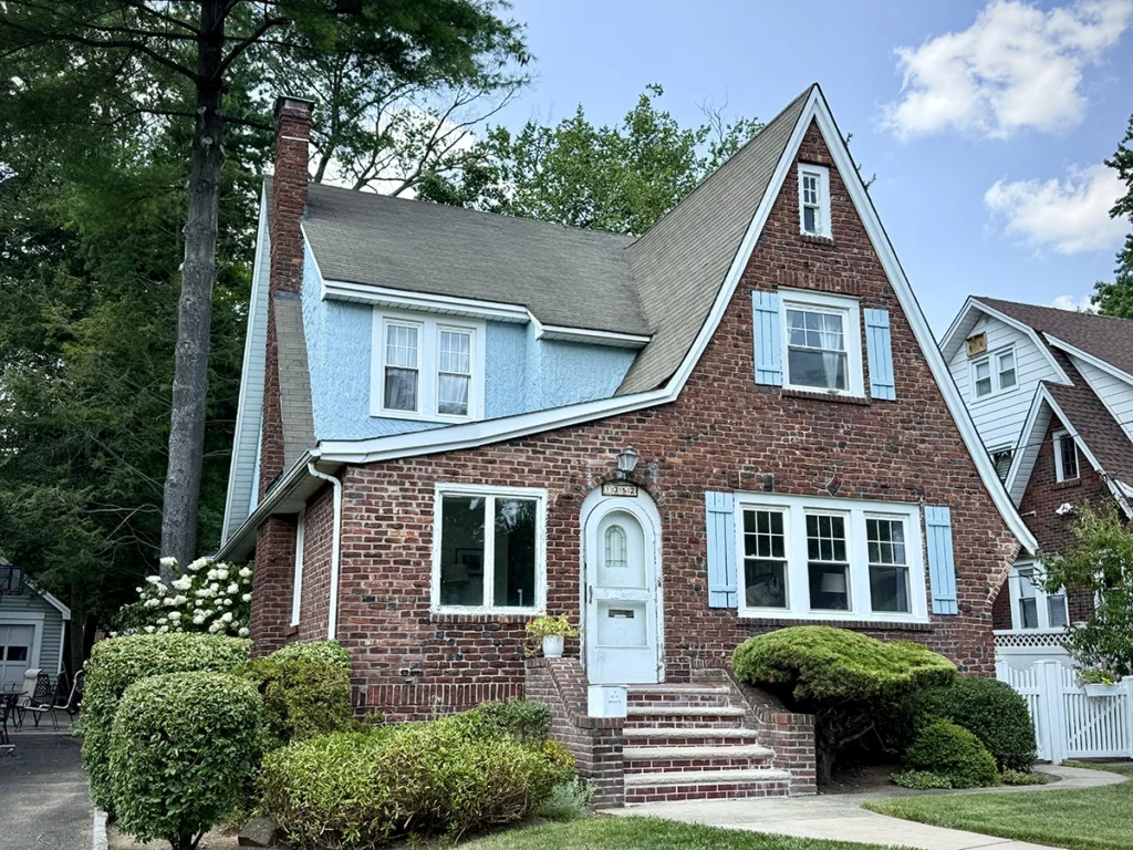 ZD Stucco Repair — Brick Tudor with blue stucco accents, Teaneck, NJ Brick Tudor-style home in Teaneck, NJ with light-blue textured stucco panels in the gables, matching shutters, and white trim; project by ZD Stucco Repair.