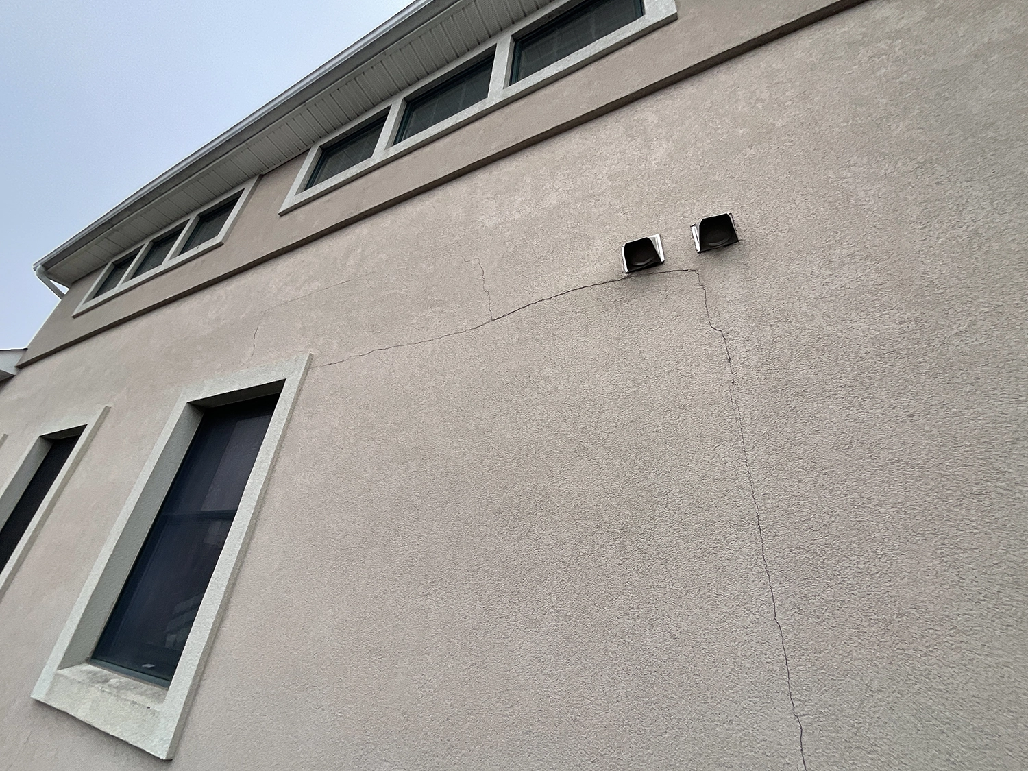 5 Common Stucco Myths Debunked – Breaking Down Popular Misconceptions 4 Wide view of a light stucco wall showing long diagonal and vertical cracks below two exhaust vents and clerestory windows.