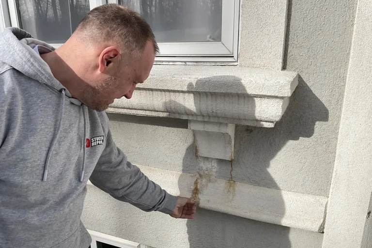 Technician points to rusty streaks and staining beneath a stucco window sill, indicating trapped moisture or failed flashing.