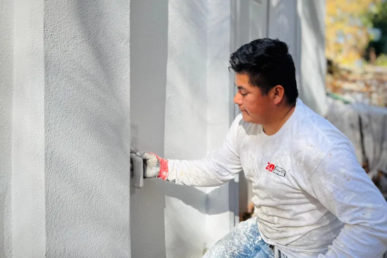 Crew member in company shirt smooths fresh stucco with a hand float on an exterior column; close-up, daytime.