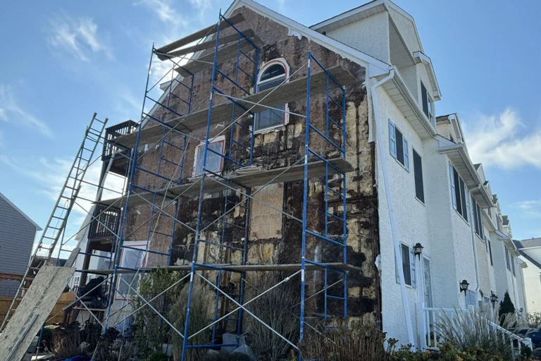 Full facade of a New Jersey home with stucco removed on scaffolding, revealing widespread wood rot and moisture damage across the entire building envelope