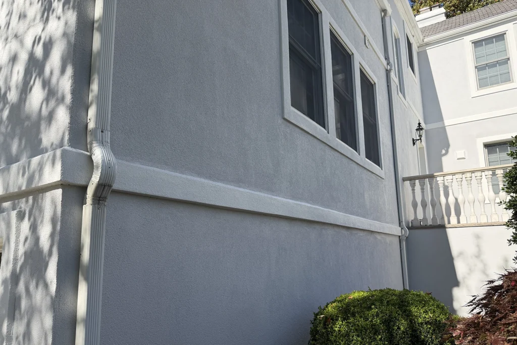 Freshly repaired gray stucco exterior wall with properly installed downspout and drainage system to prevent future water infiltration and wood rot in New Jersey