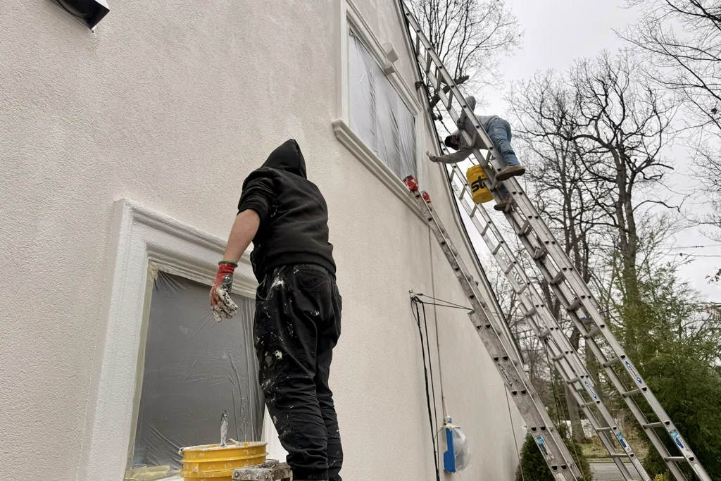 Two contractors working from ladders while applying a fresh white coating to a stucco house
