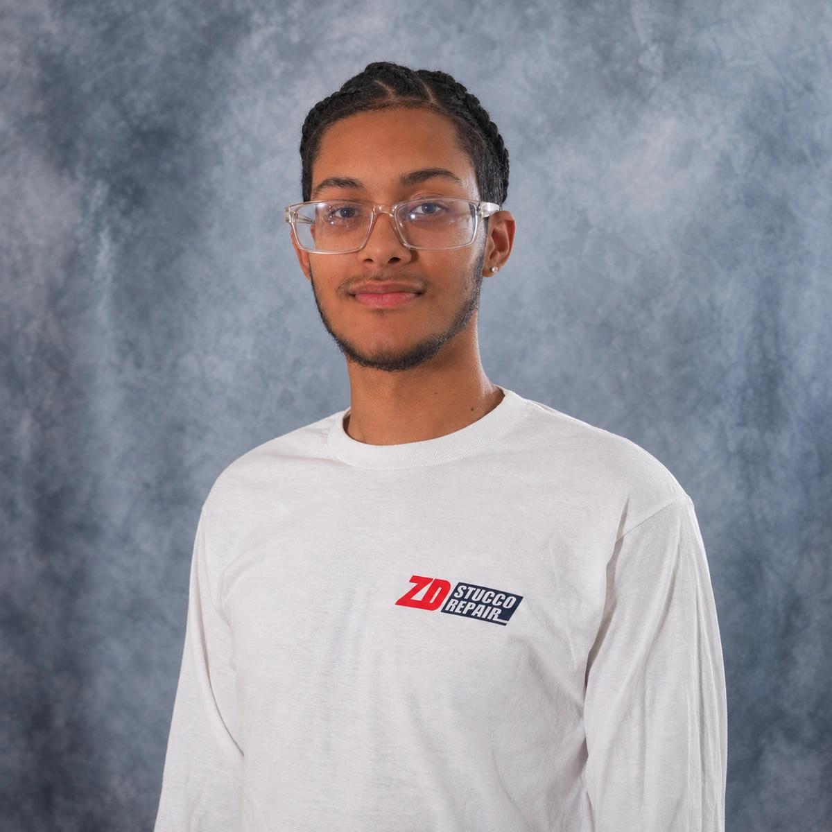 Alex, Warehouse Manager at ZD Stucco Repair, wearing clear glasses and a white company shirt against a gray studio backdrop.